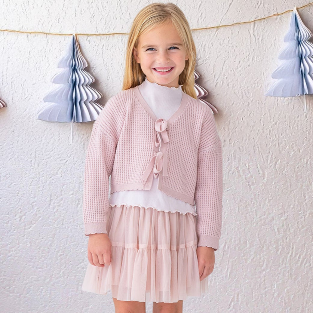 Young girl in a pink dress standing in front of a white wall with decorative trees.