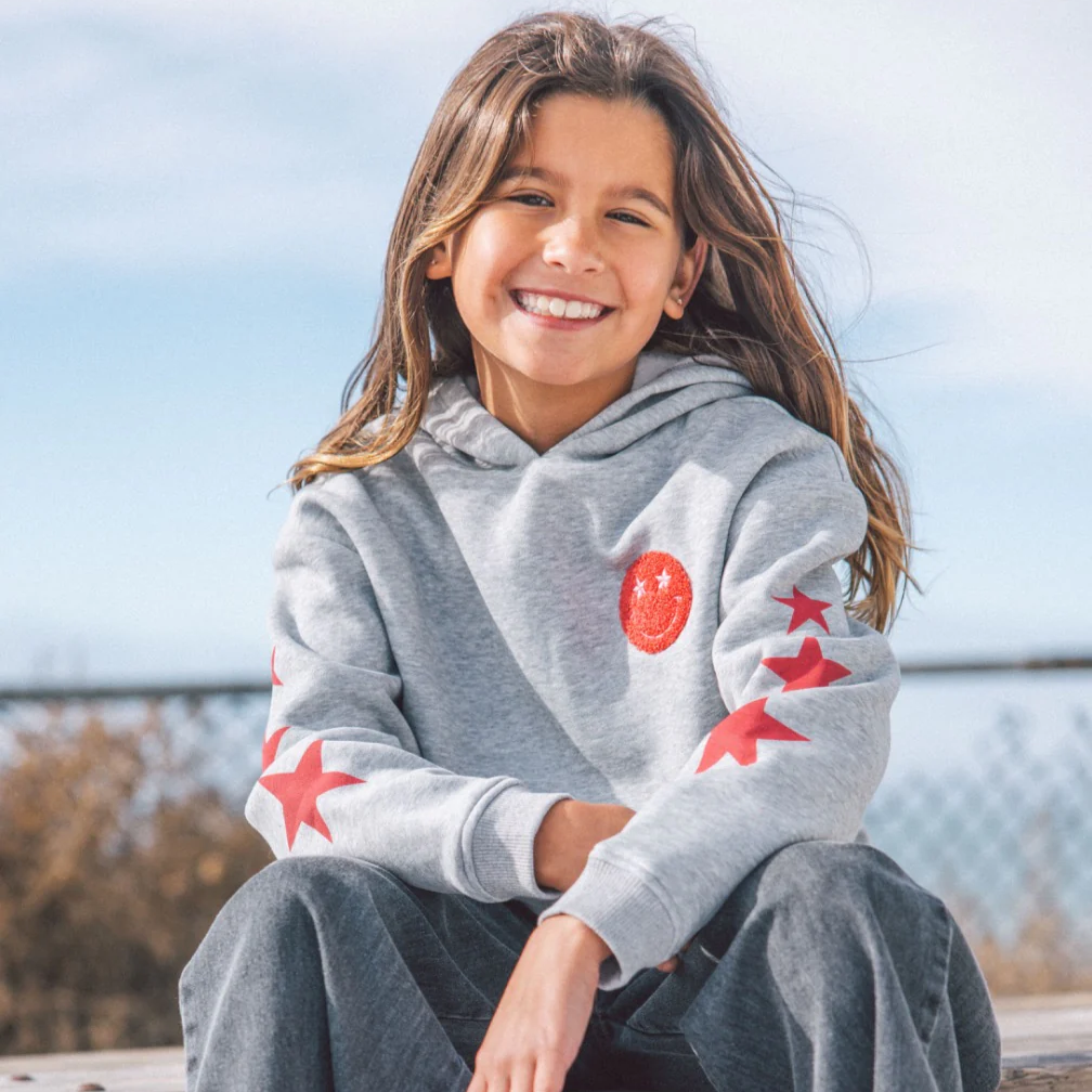 Young girl wearing a gray hoodie with red star designs, sitting outdoors.