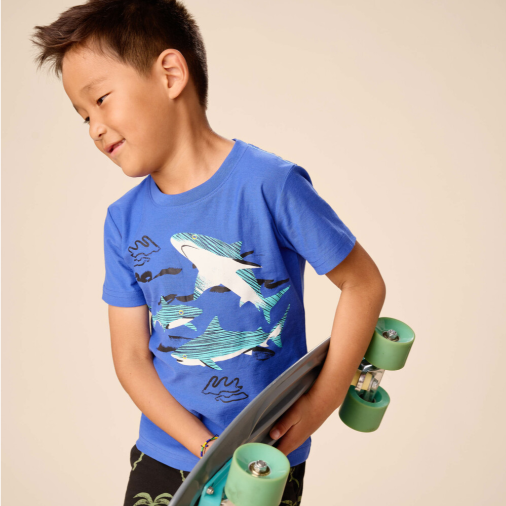 Child wearing a blue t-shirt with shark design holding a skateboard against a beige background