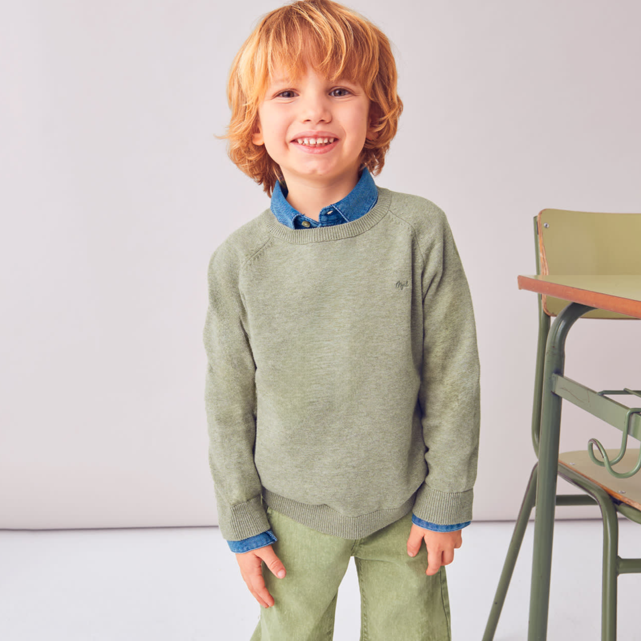 Child wearing a green sweater and pants standing indoors with a chair in the background.