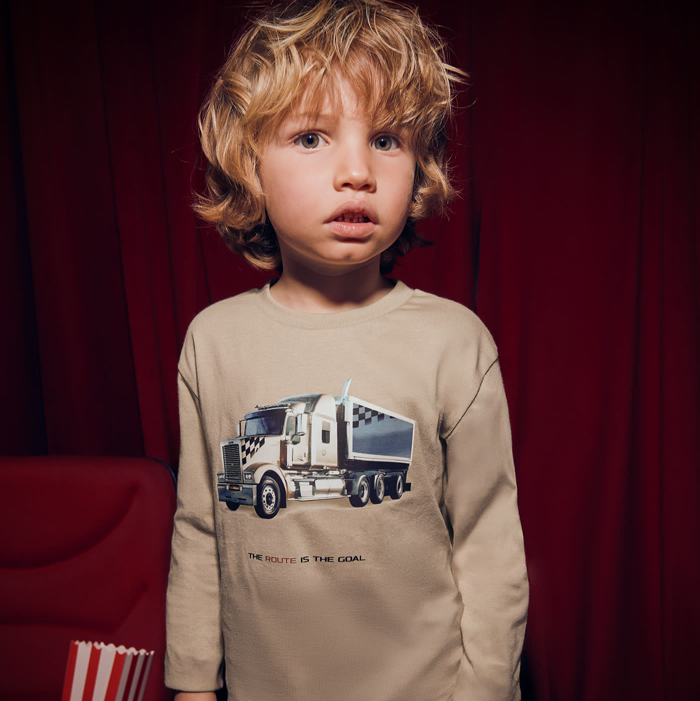 Child wearing a beige long-sleeve shirt with a truck graphic against a red curtain background