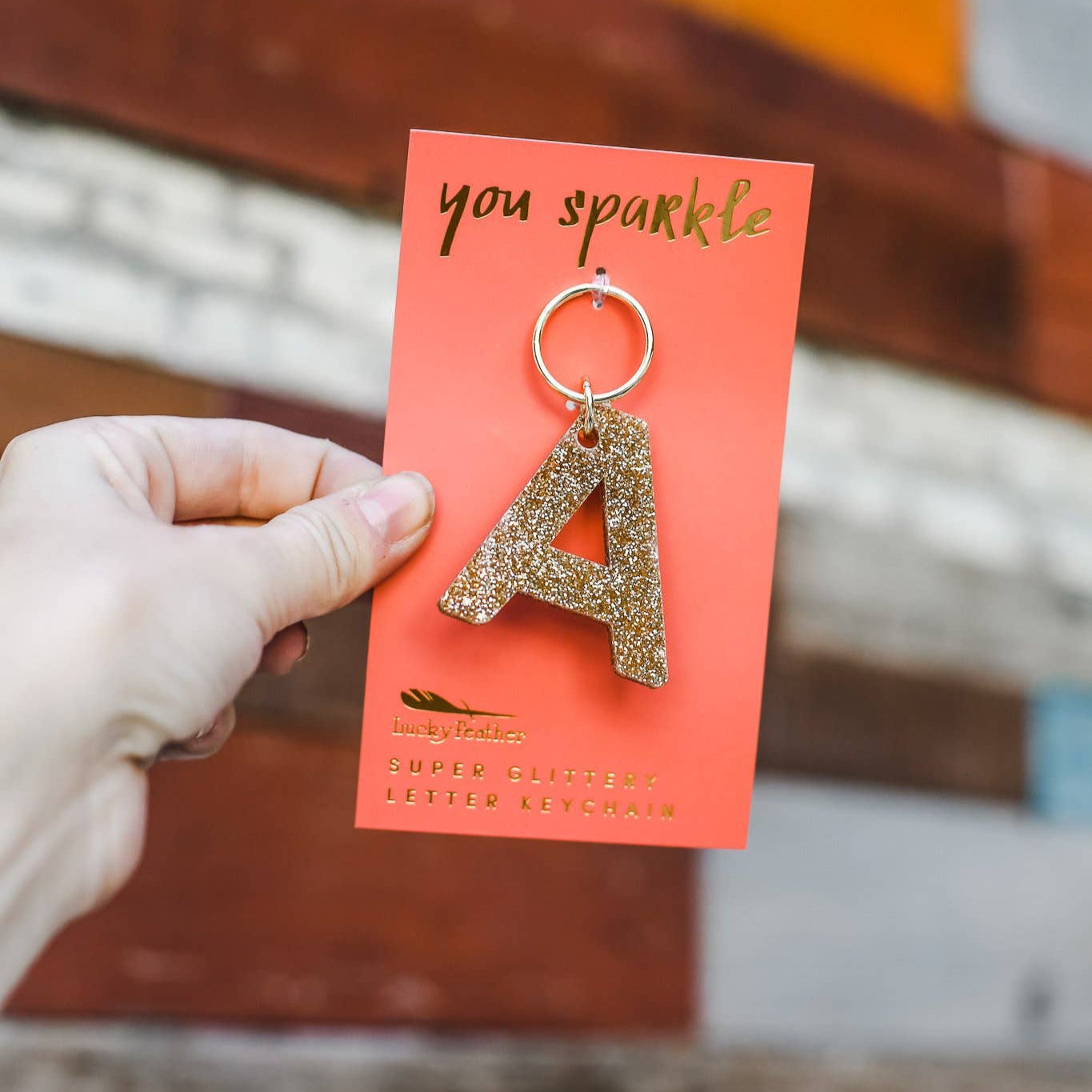 Gold letter 'A' keychain on a red card with 'you sparkle' text, held against a blurred background.