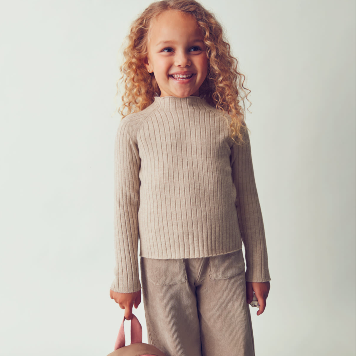 Child in beige outfit with a small backpack on a light background