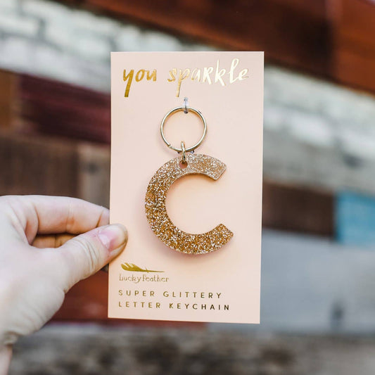 Gold glittery letter 'C' keychain on a pink card held by a hand with a blurred background
