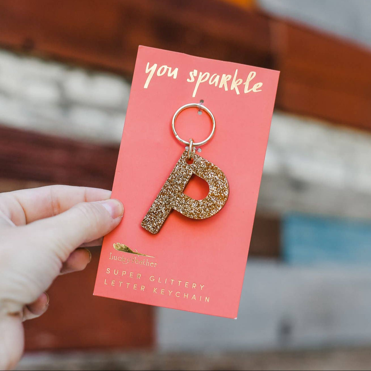 Gold letter 'P' keychain on a pink card with 'you sparkle' text, held by a hand.