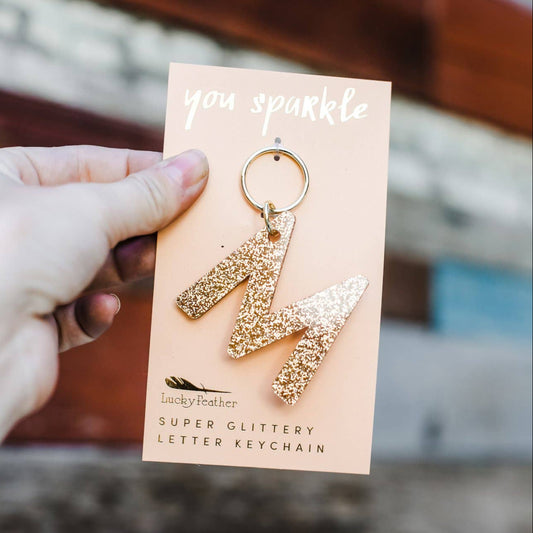 Gold glittery letter 'M' keychain on a card held by a hand with a blurred background
