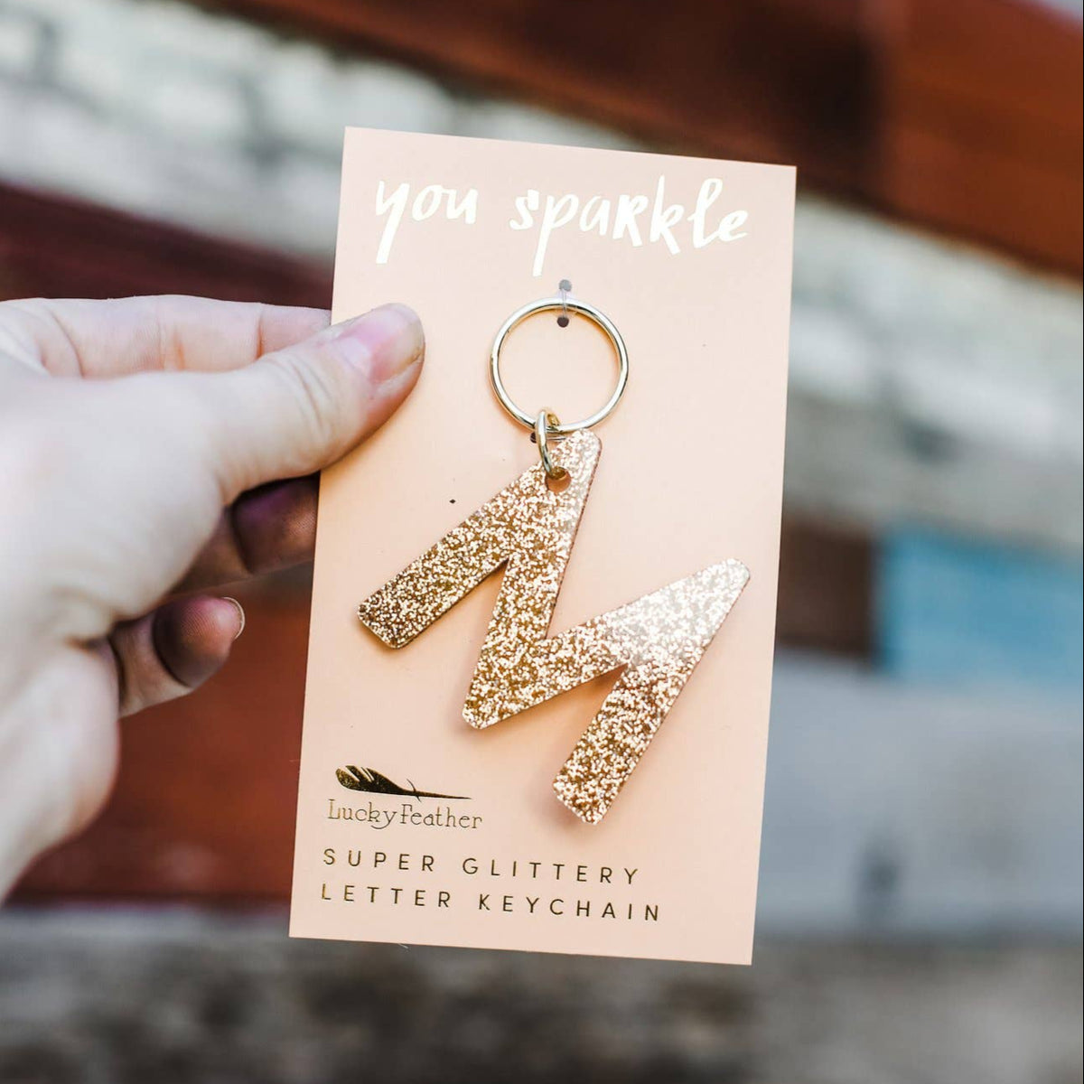 Gold glittery letter 'M' keychain on a card held by a hand with a blurred background