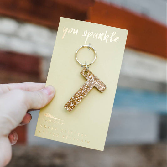 Gold letter 'T' keychain on a card with 'you sparkle' text, held by a hand against a blurred background.