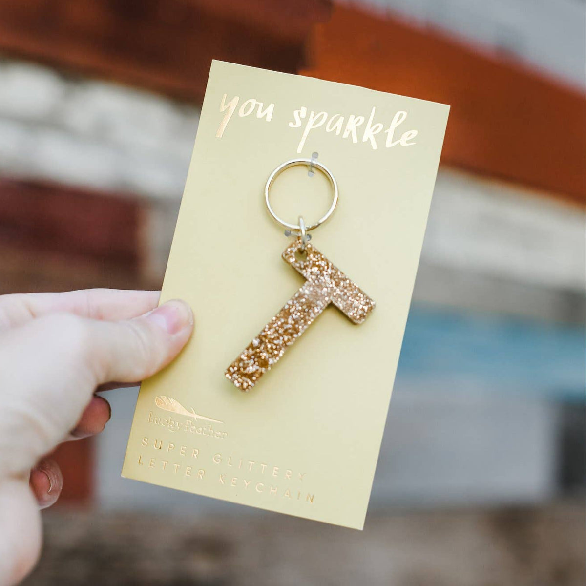 Gold letter 'T' keychain on a card with 'you sparkle' text, held by a hand against a blurred background.
