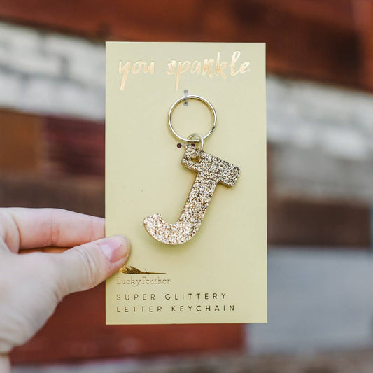 Gold glittery letter 'J' keychain on a card held by a hand with a blurred background