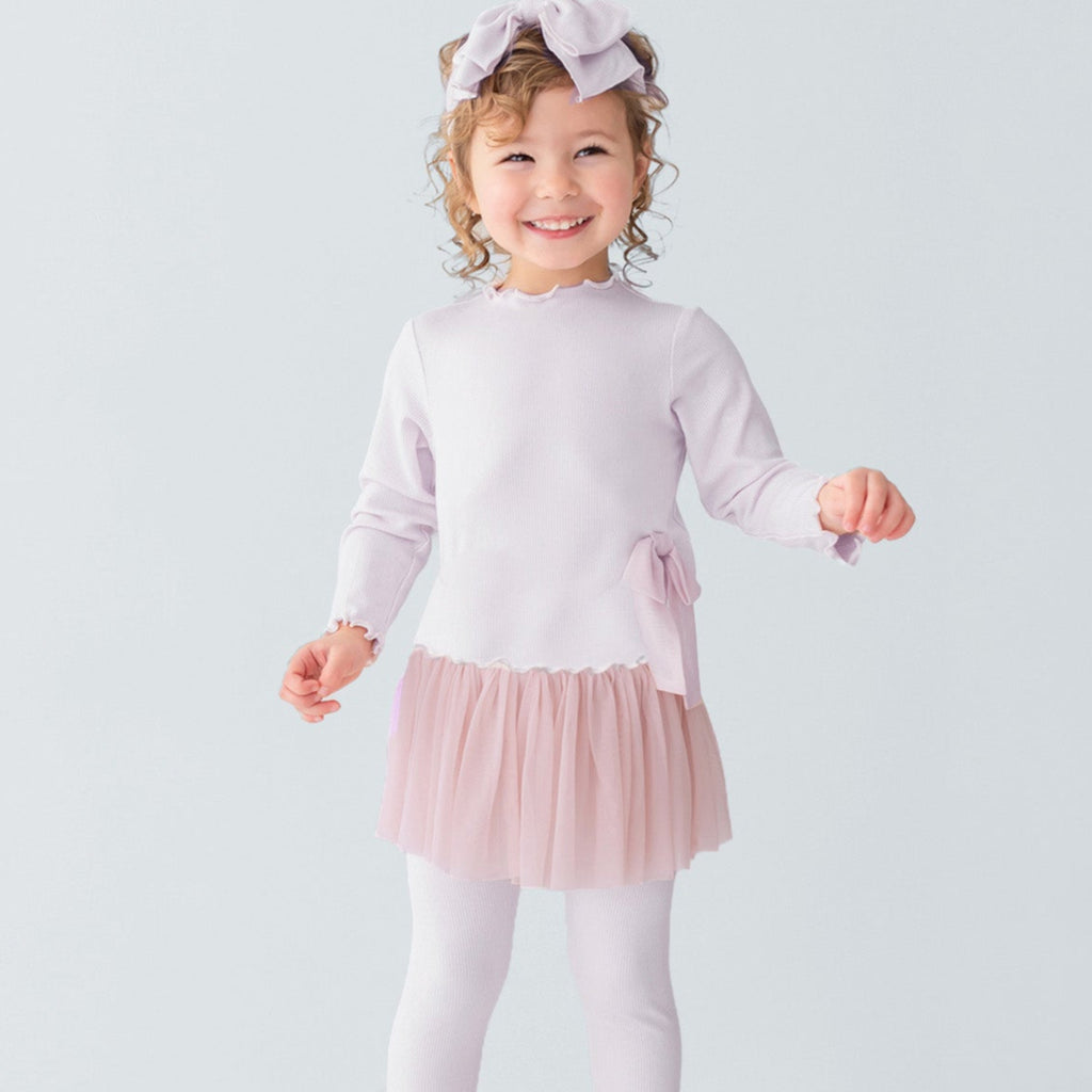 Young girl in a light pink dress with a tutu skirt and white leggings standing on a white platform.