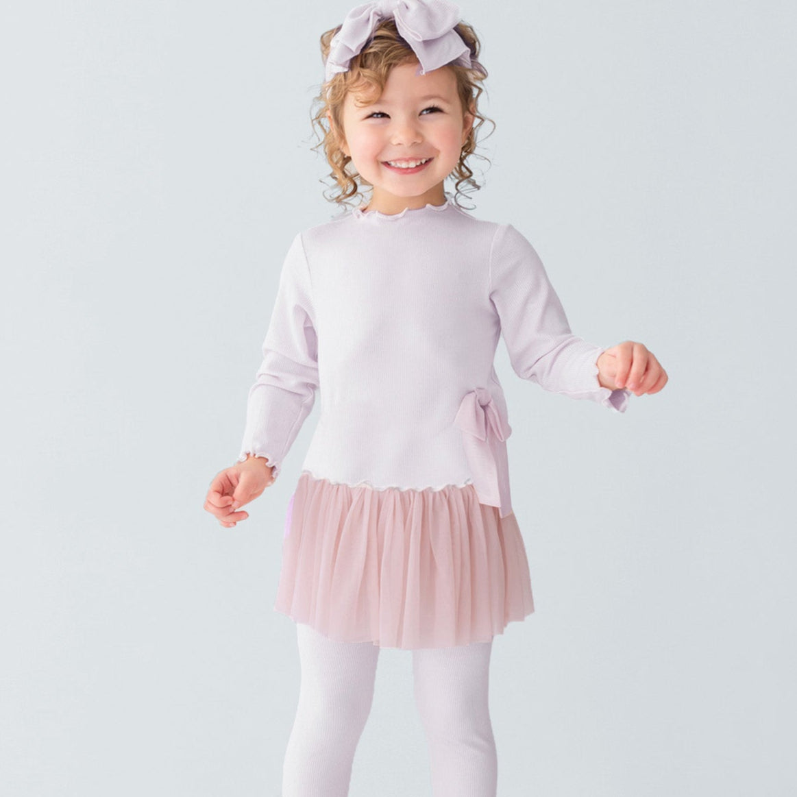 Young girl in a light pink dress with a tutu skirt and white leggings standing on a white platform.