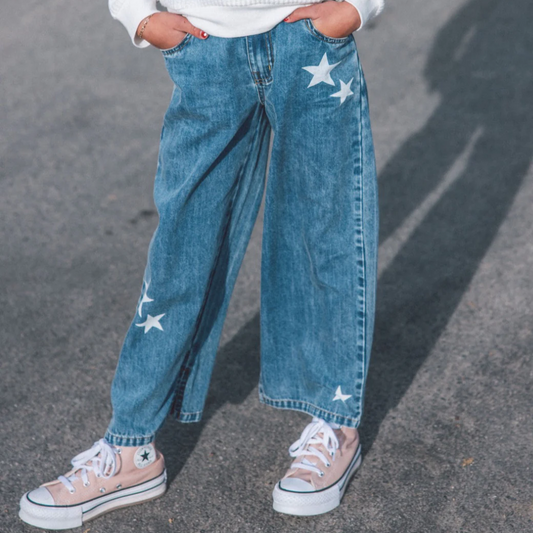 Person wearing blue jeans with star patches and white sneakers on a gray pavement.