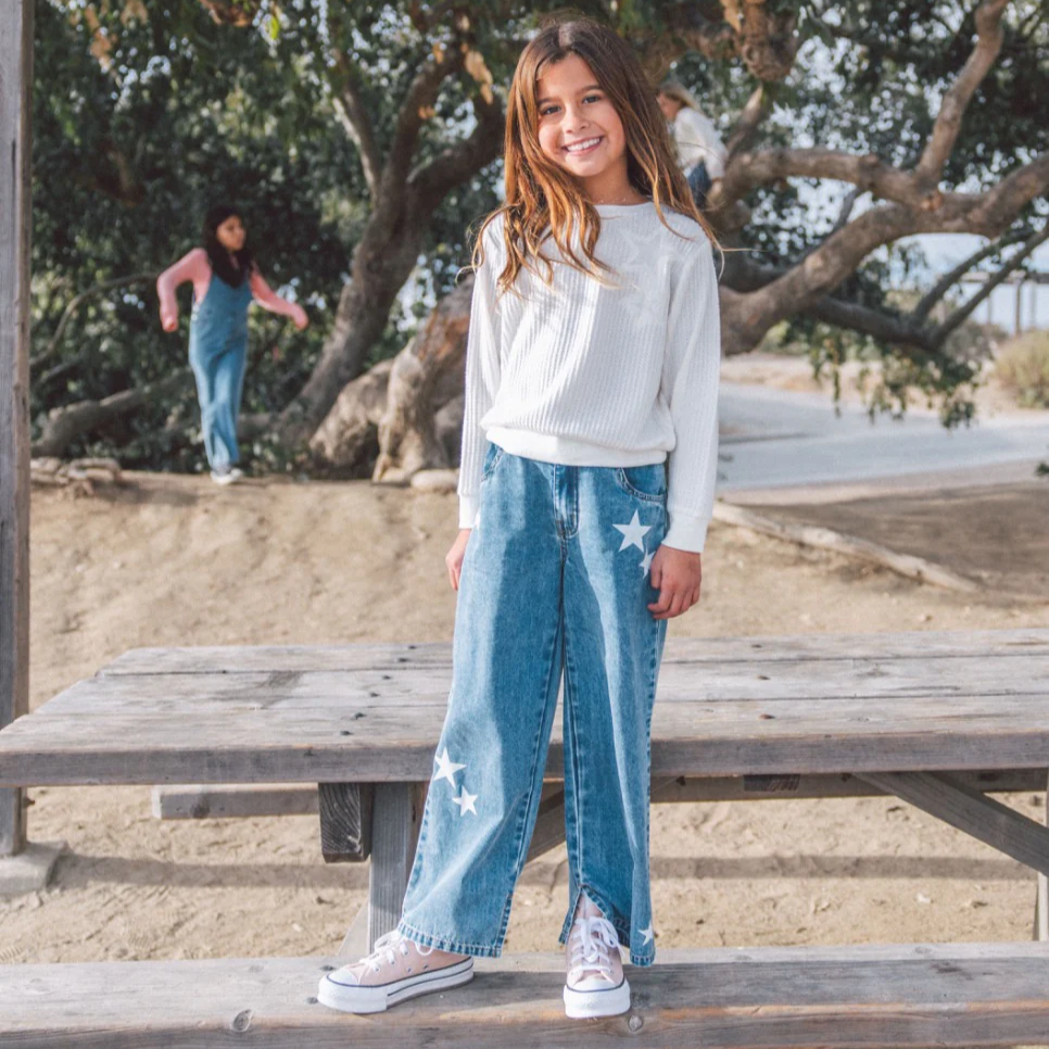 Young girl standing on a wooden bench at a park with another person in the background.