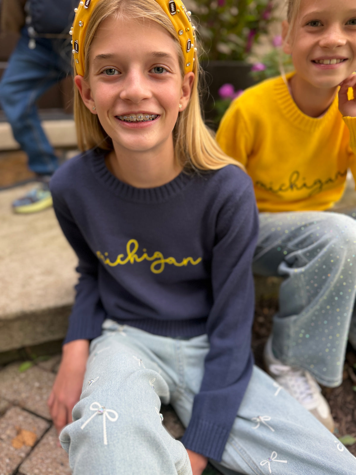A young girl wearing a navy blue crewneck sweater with 'michigan' written in yellow script across the chest, paired with light blue bow-patterned jeans and a yellow patterned headband. She is smiling and sitting outdoors, with another child in a yellow sweater visible in the background.