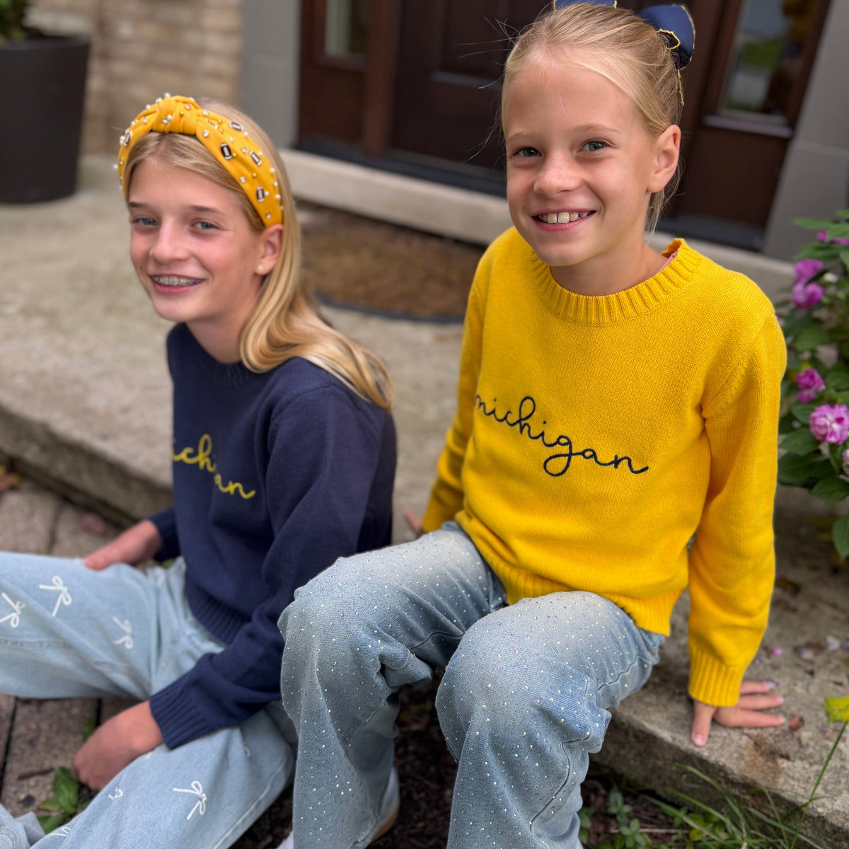 Two young girls sitting side-by-side outdoors in front of a house. The girl on the right wears a bright yellow knit sweater with 'michigan' embroidered in black script, paired with light blue glittery jeans.