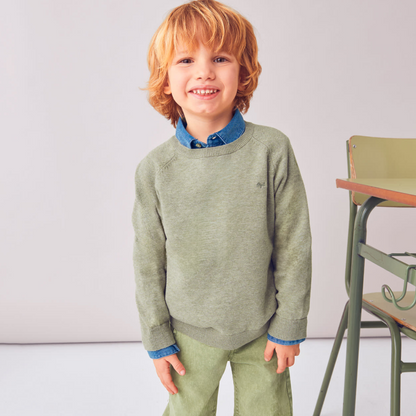 Child wearing a green sweater and pants standing indoors with a chair in the background.