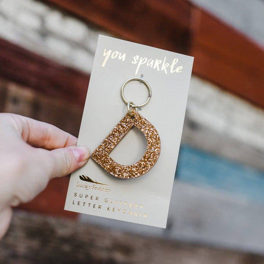 Gold letter 'D' keychain on a card with 'you sparkle' text, held against a blurred background.