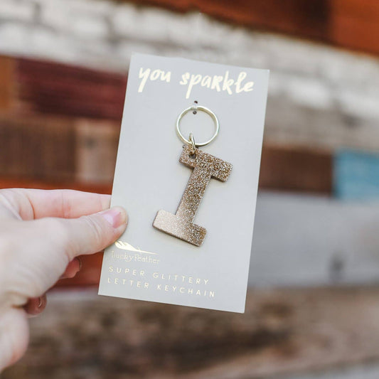 Gold letter 'I' earring on a card with 'you sparkle' text, held by a hand against a blurred background.
