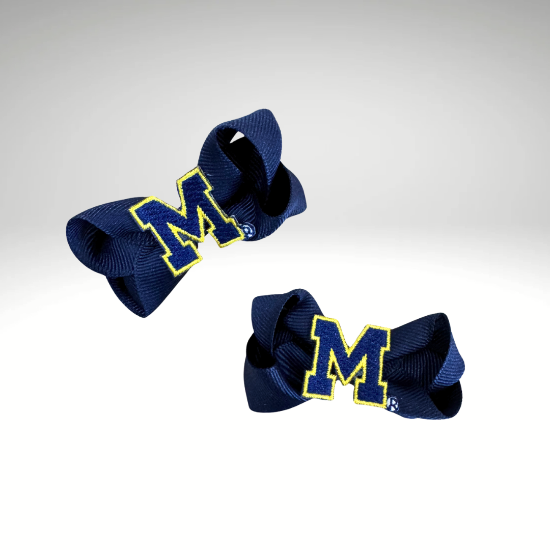Two navy blue grosgrain ribbon hair bows with yellow and white embroidered letter 'M' accents, displayed on a light gray gradient background.